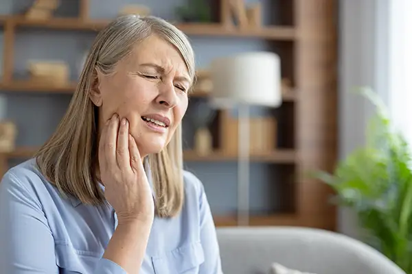 Elderly woman sitting at home, holding her jaw and grimacing in pain due to pain from a dental crown.