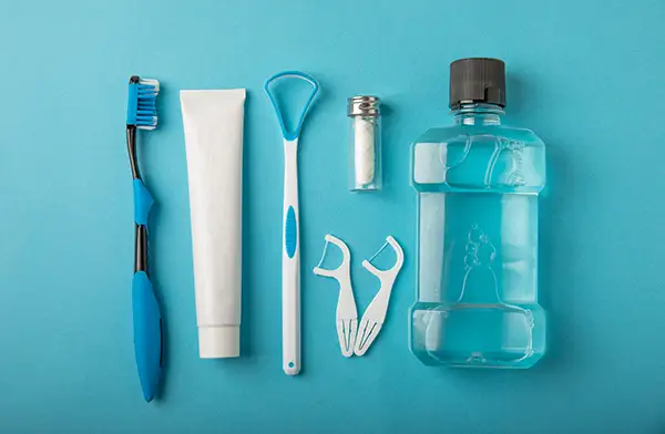 Oral hygiene products, including a toothbrush, toothpaste, tongue scraper, floss, and mouthwash, arranged neatly on a blue background.